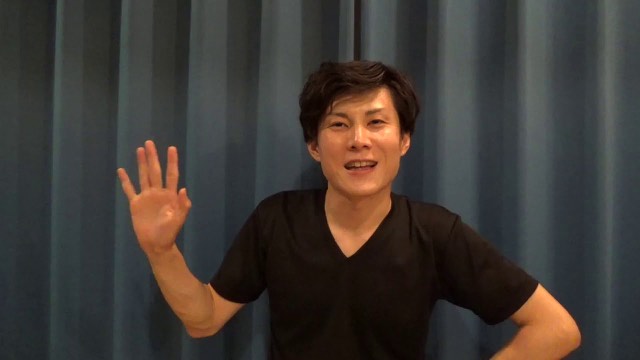 青いカーテン背景で黒Tシャツ姿の本人が手を挙げて笑う様子(動画「トラブルを人のせいにしたら赤信号です 不幸になります」より)