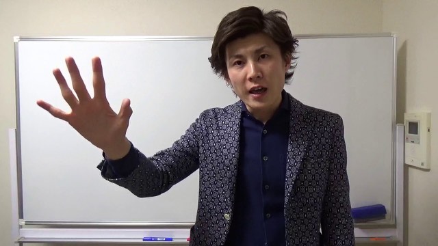ホワイトボード前で片手を挙げる柄物ジャケットの本人(動画「月収30~60万のスロプロから抜け出すには…」より)